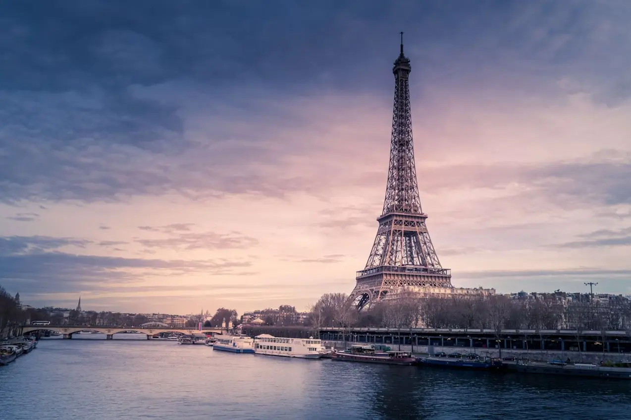 Une vue sereine sur la Tour Eiffel au crépuscule, avec un ciel rose pourpre et des nuages au-dessus. La tour se dresse élégamment sur le côté droit, tandis que la calme rivière Seine coule en dessous, reflétant les teintes pastel. Les bateaux sont amarrés le long de la rivière, incarnant le charme recherché par les chasseurs d'appartement Paris 7.