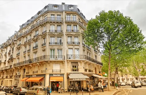 Un coin de rue parisien du 16ème arrondissement abrite un immeuble classique de style haussmannien avec des balcons en fer forgé et de nombreuses fenêtres. Le rez-de-chaussée accueille des cafés et des boutiques aux auvents orange. A proximité, des voitures garées et un grand arbre feuillu sont visibles sous un ciel nuageux, parfait pour tout chasseur immobilier Paris 16.