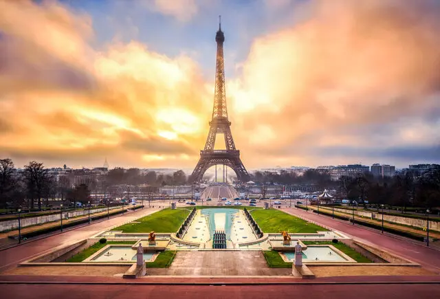 Une vue imprenable sur la Tour Eiffel à Paris au coucher du soleil. Le ciel est rempli de teintes orange et jaune vibrantes, projetant une lueur chaleureuse sur la scène. Le premier plan présente des jardins paysagers et des fontaines, avec des allées menant à la structure emblématique, idéale pour un Chasseur immobilier à Paris 16.